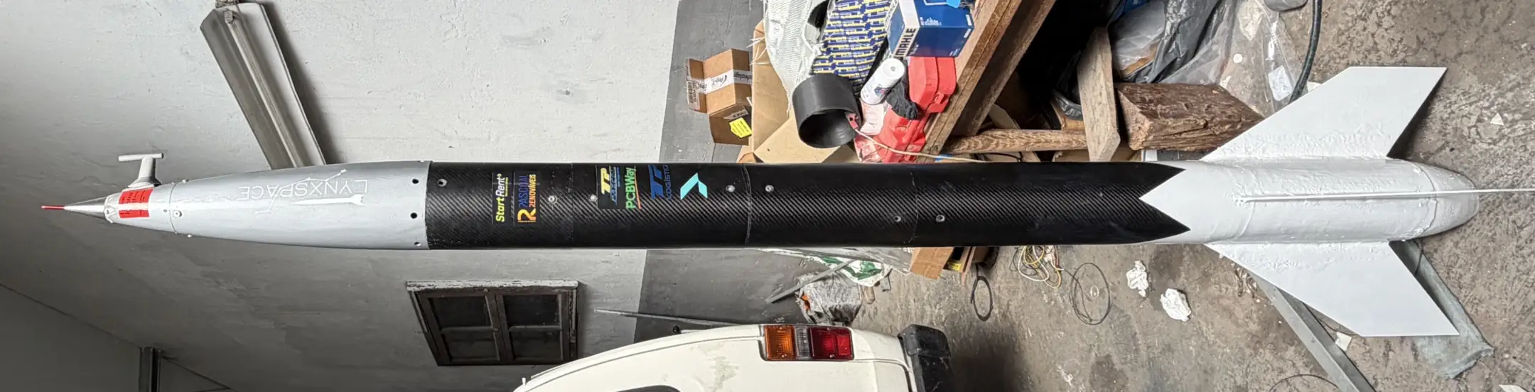 A fuselagem completa do Lynxrocket V5 na oficina: ogiva em fibra de vidro com ponta de tampa vermelha e sonda pitot-estática, tubo de corpo em fibra de carbono com os autocolantes dos parceiros e o conjunto de quatro aletas em carbono