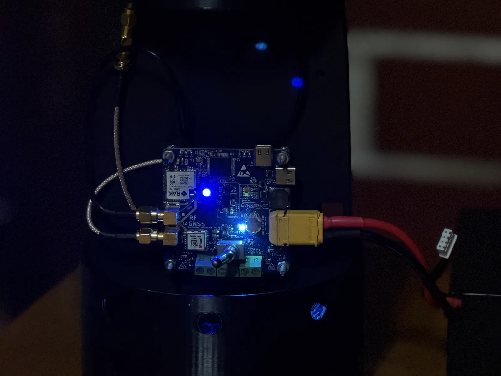 LynxYNav V6.1 ligado no seu suporte de aviónica dentro do acoplador da fuselagem: LEDs de estado azuis acesos, cabo de alimentação XT60 ligado, dois cabos de antena SMA e a antena LoRa por cima