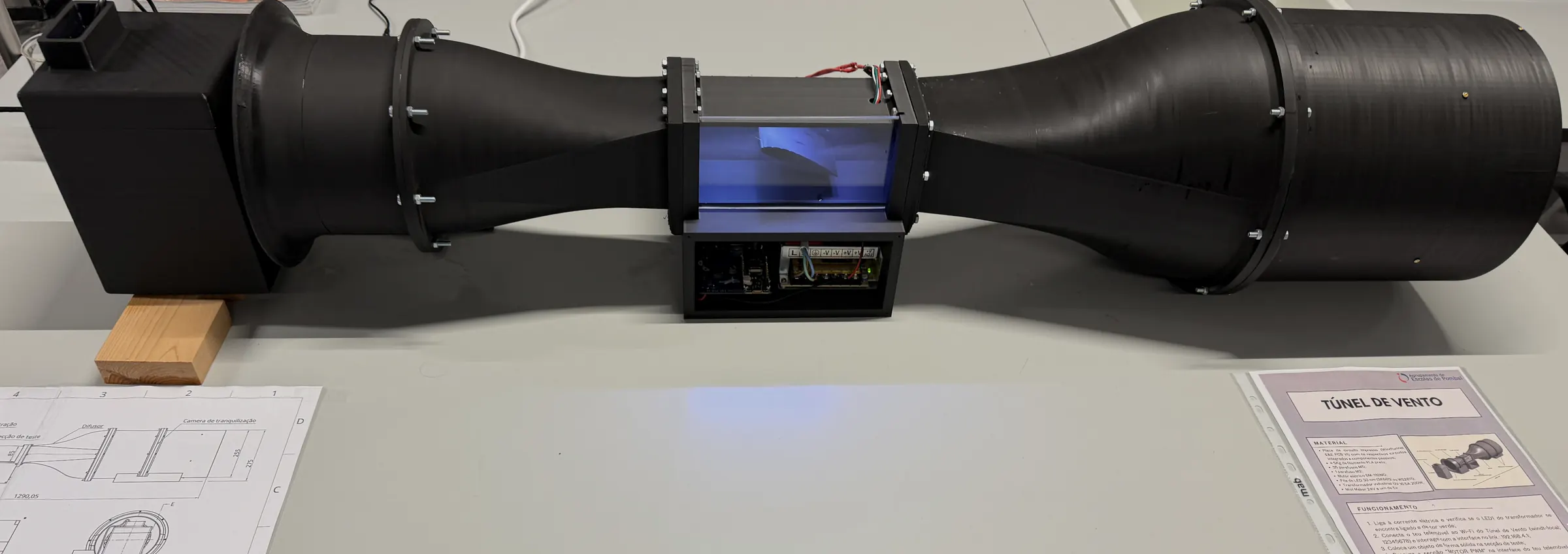 O túnel de vento completo numa bancada de laboratório, impresso a preto: o reservatório de vapor e a entrada em boca de sino à esquerda, o cone de contração, a secção de ensaio iluminada com o compartimento de eletrónica por baixo, o difusor e a câmara de saída à direita. O desenho de conjunto impresso está pousado na bancada, à esquerda